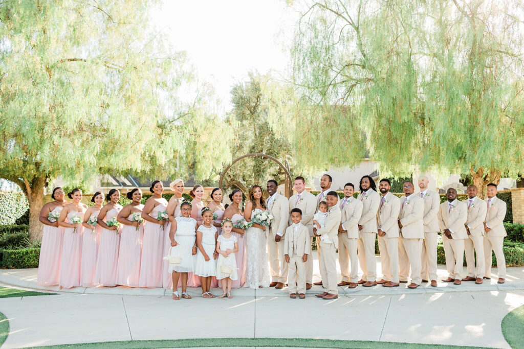 Bridal party with bride and groom posing for pictures in front of luscious weeping willow trees at rustic wedding.